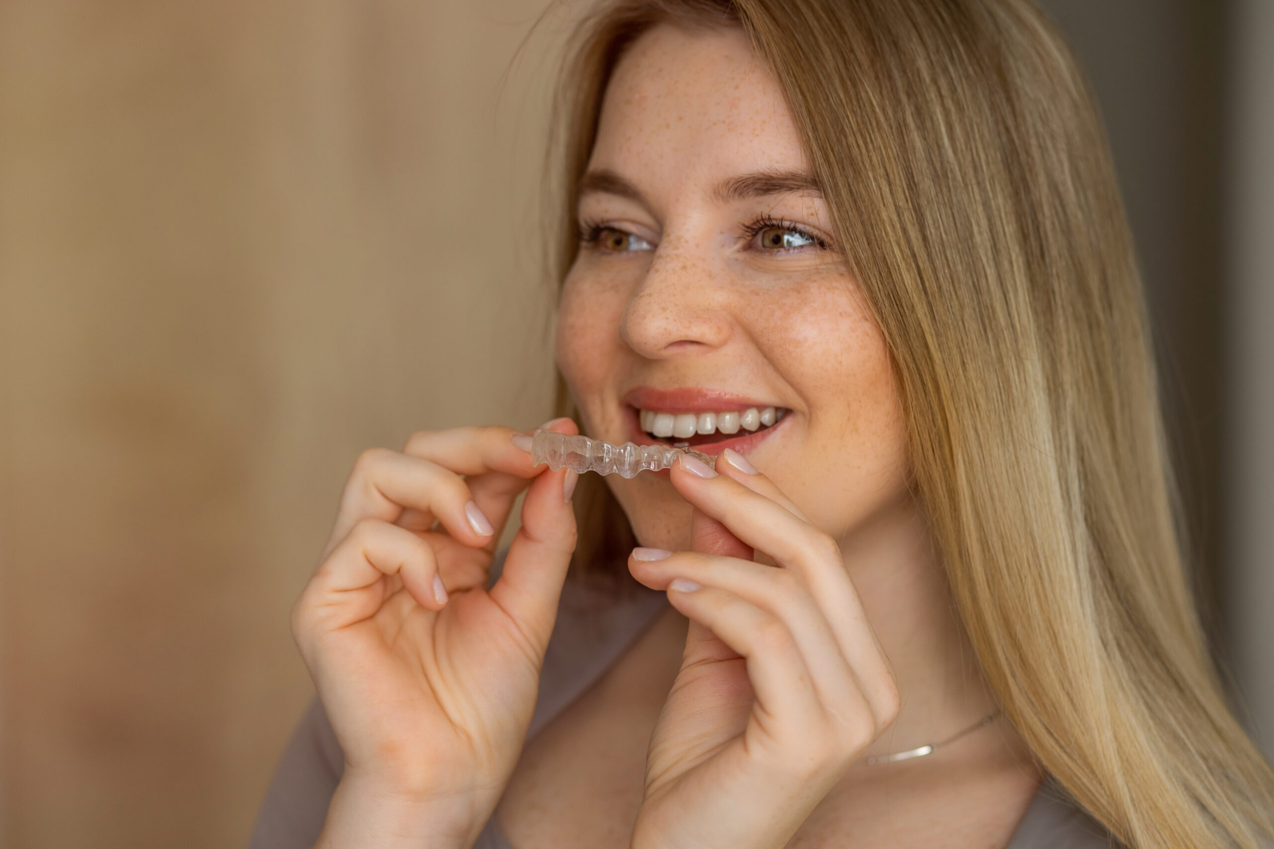 Smiling woman holding transparent aligner as orthodontic dental tool A young woman smiles while holding a clear dental aligner, showcasing modern orthodontic care. Perfect teeth correction and aesthetic restoration aid concept.