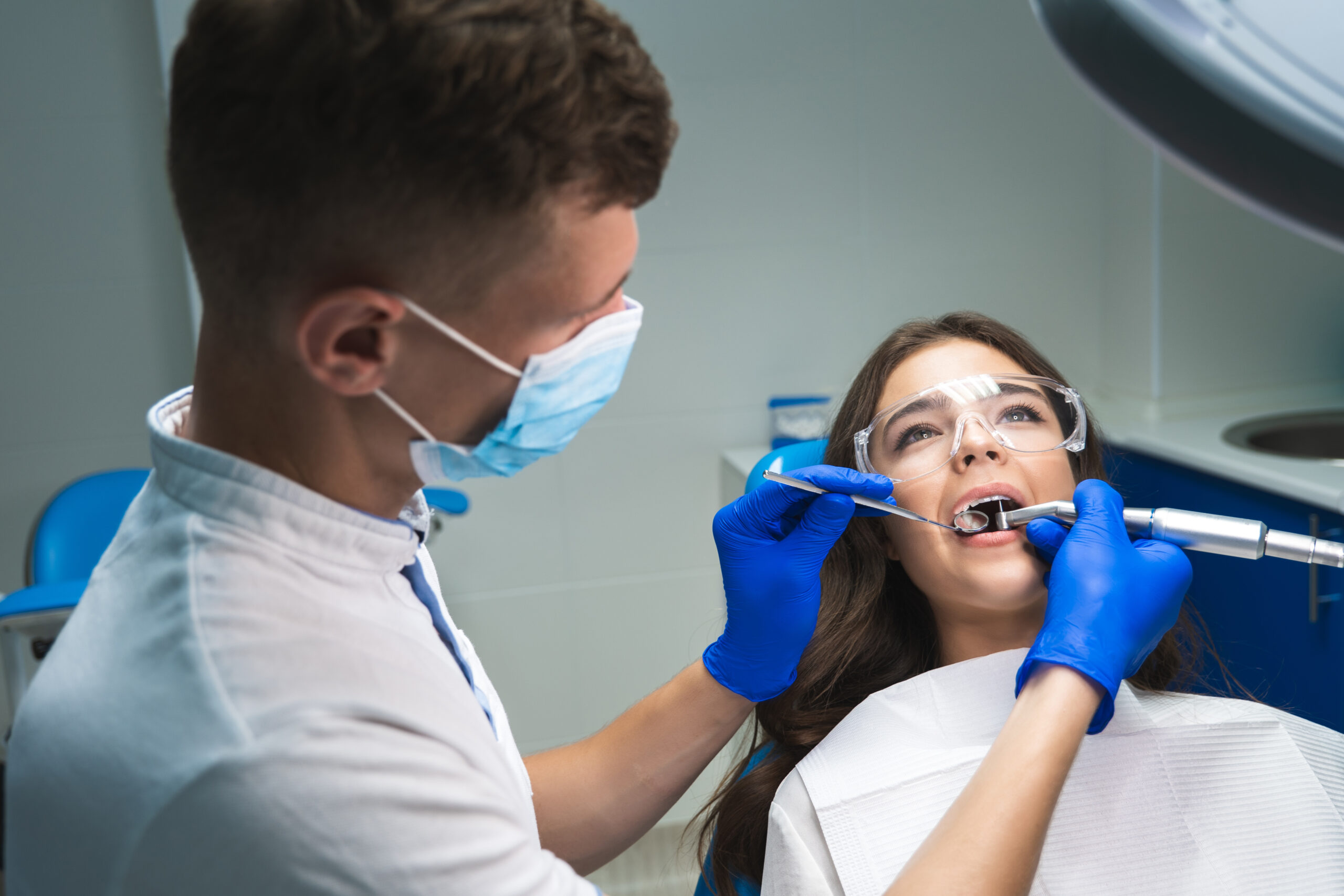 dentist in medical mask filling the patient's root canal while she is in dental chair wearing safety glasses under the medical lamp in clinic.