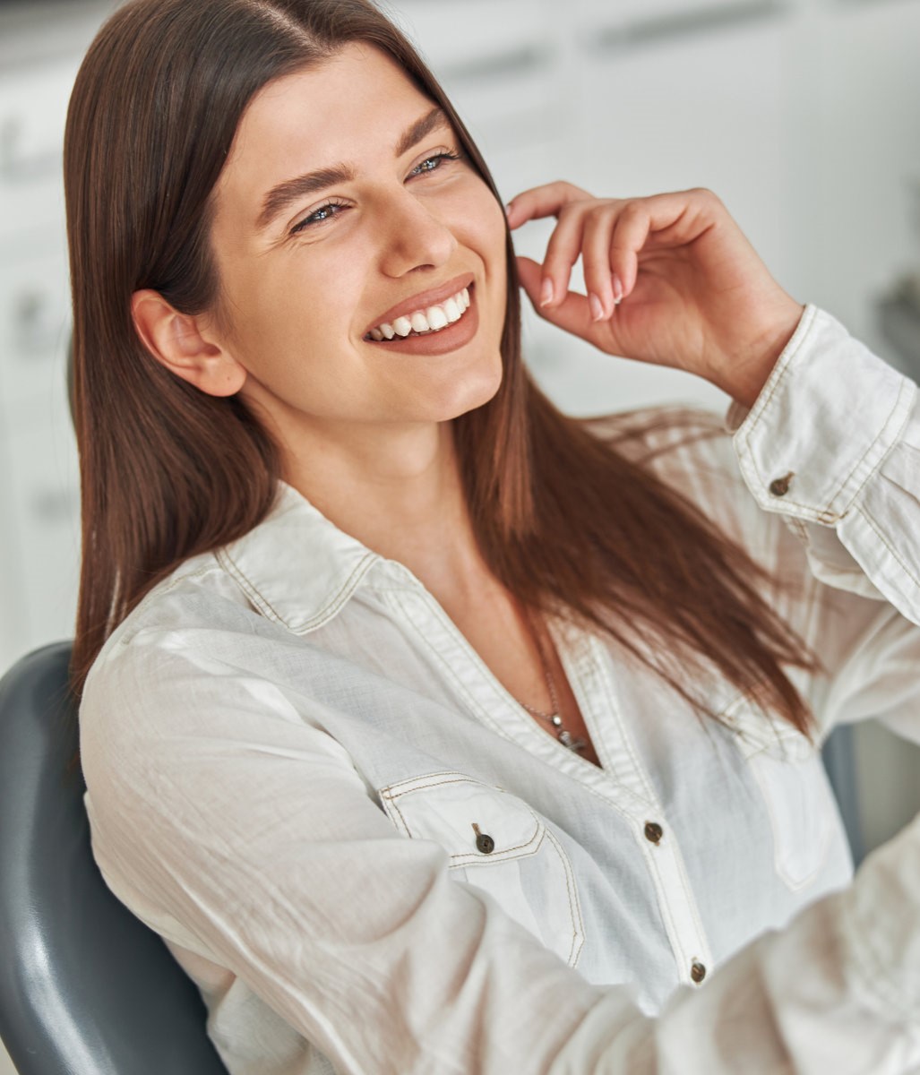 AdobeStock_428235579_result1 Happy young woman smiling checking out her perfect healthy teeth in the mirror, sitting in a dental chair at the dentist office