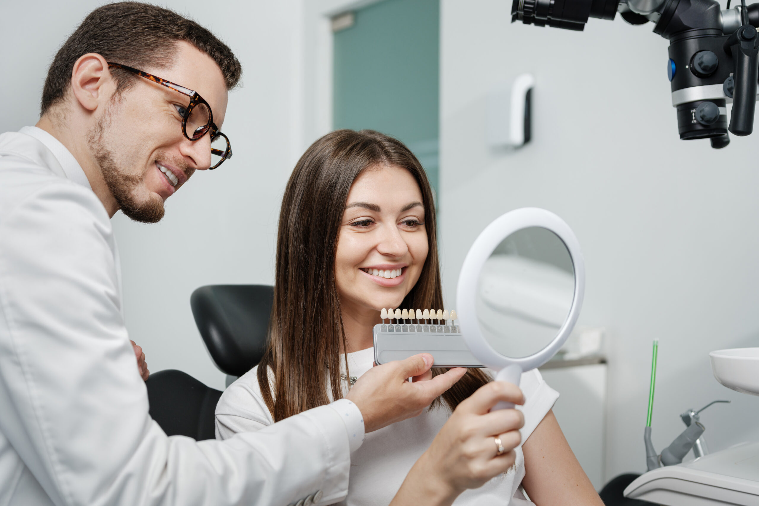 Young male dentist in white coat show veneers enamel color palette for beautiful patient woman sitting in a dental unit. Healthcare oral aesthetic treatment.