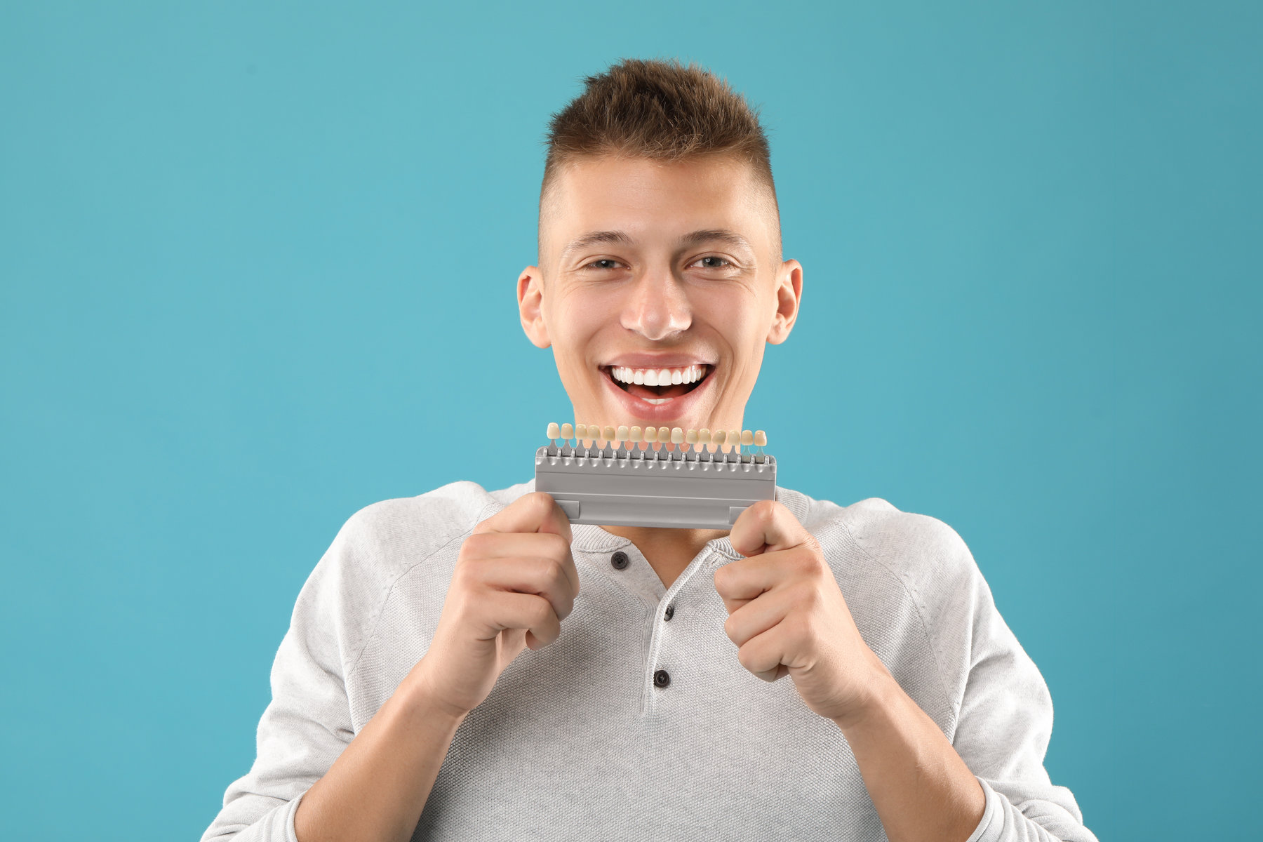 Happy young man with teeth color samples on light blue background. Dental veneers Happy young man with teeth color samples on light blue background. Dental veneers
