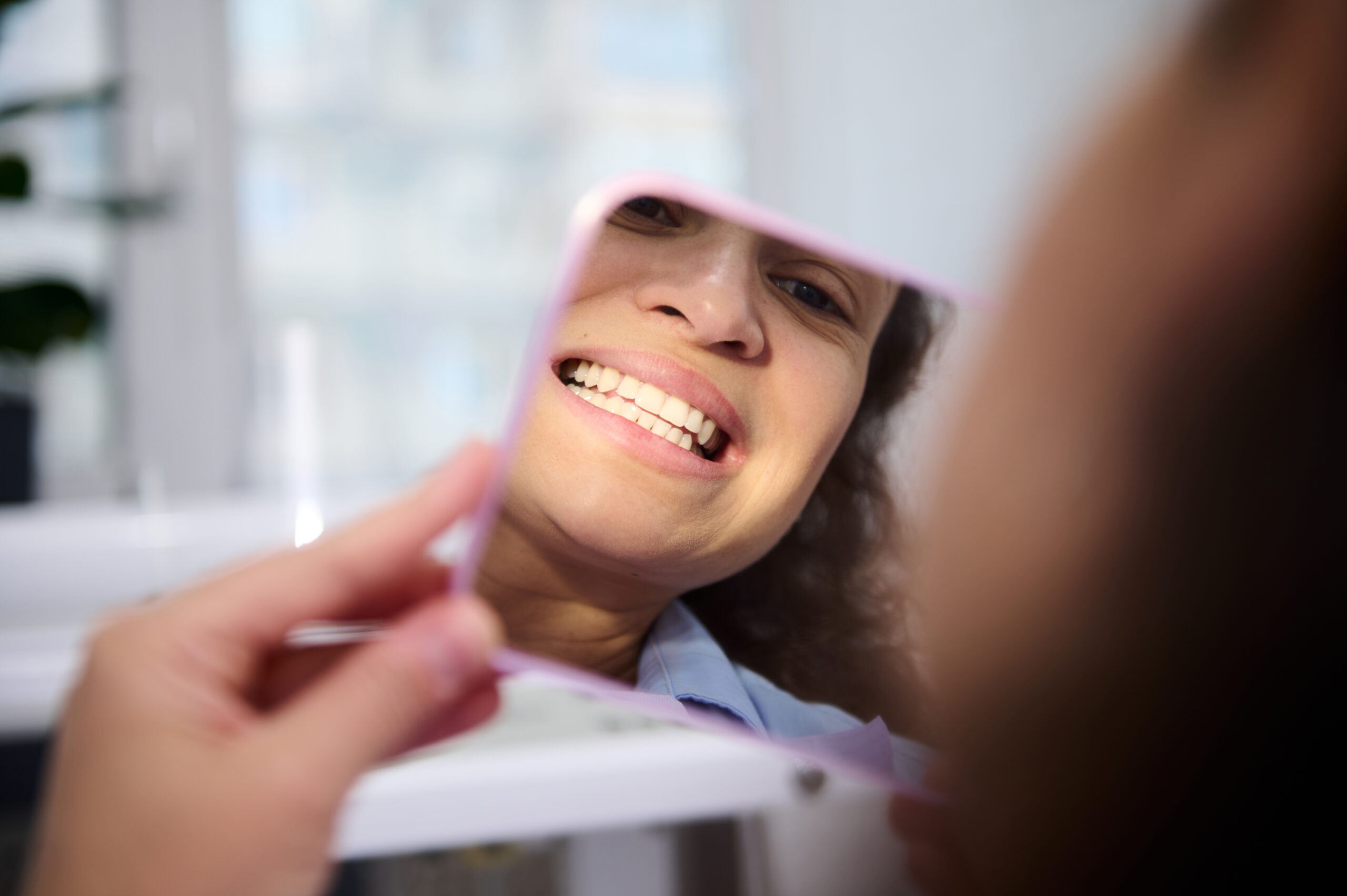 Reflection in mirror of a female patient in dental chair, admiring her smile and teeth after teeth bleaching procedure