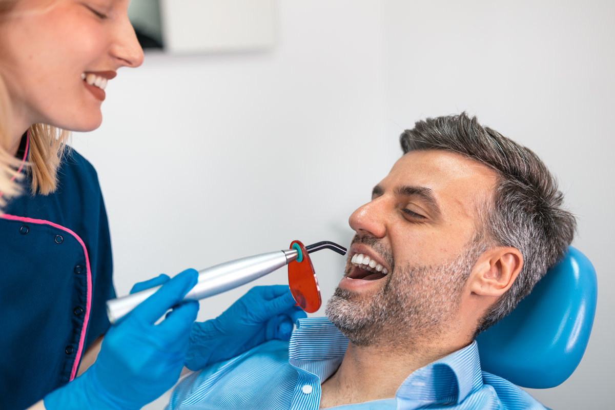 Dental curing light treatment. Professional female dentist using UV lamp to harden filling on a male patient in clinic, Healthcare and Medicine concept. Modern dentistry procedure for tooth repair.